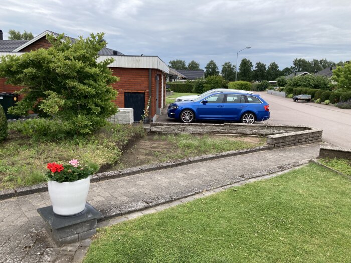 Parkeringsplats med en blå bil parkerad framför ett tegelhus, omgiven av häckar och träd, en vit blomkruka med blommor i förgrunden.