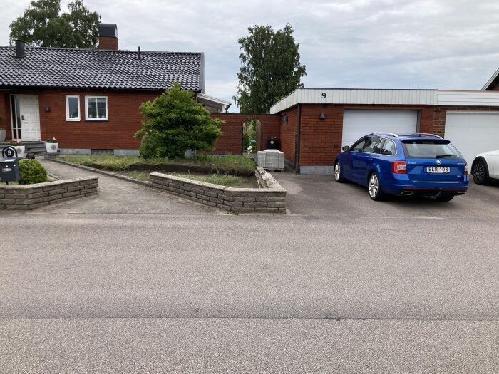 En röd tegelvilla med en parkering framför, en blå bil står parkerad. Framtiden av huset med murkanter har blivit omgjord och marken bearbetad.