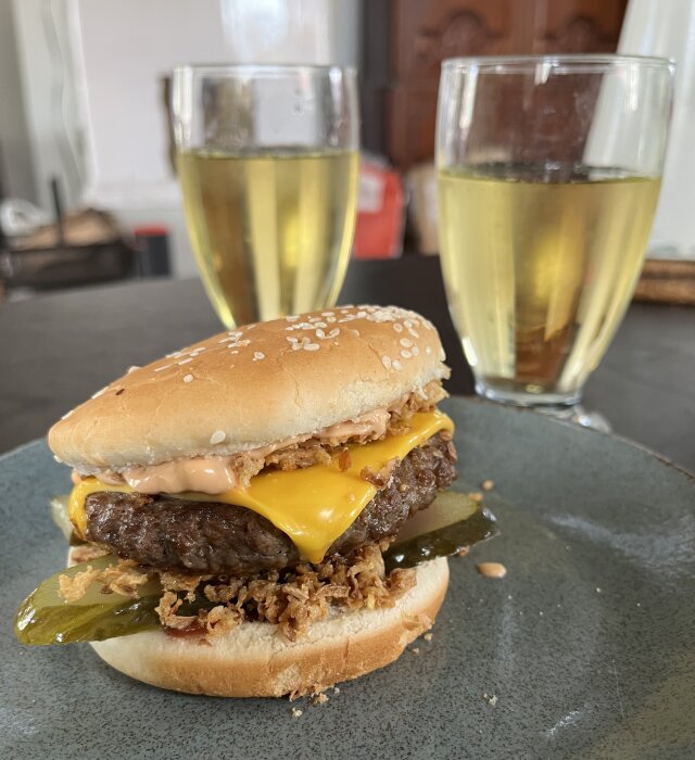 Hamburgare med ost, gurka, dressing och rostad lök på en tallrik. I bakgrunden två glas dryck på ett bord.