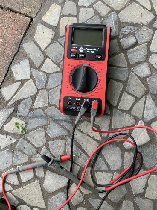 Digital multimeter av märket Powerfix KH 3320 med kablar liggande på stenbeläggning.