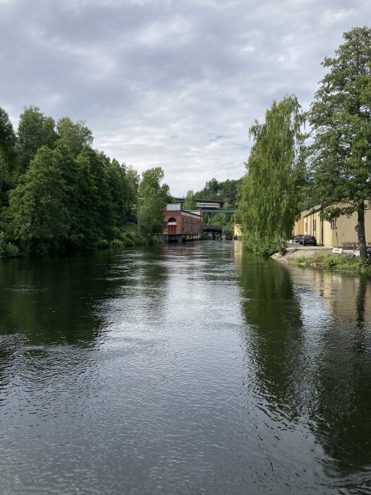 En stilla flod med gröna träd längs kanterna, en röd byggnad i bakgrunden och en gul byggnad till höger under molnig himmel.