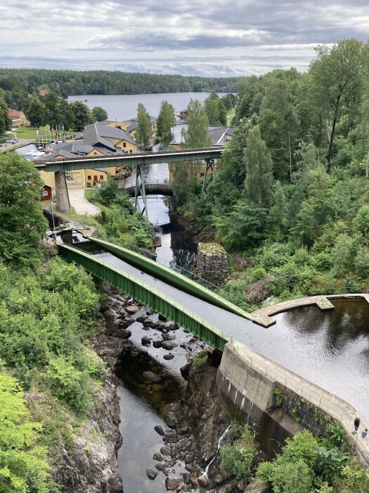 Vy över en gammal industrimiljö med byggnader längs ett vattendrag, med gröna träbroar, skog och en sjö i bakgrunden.