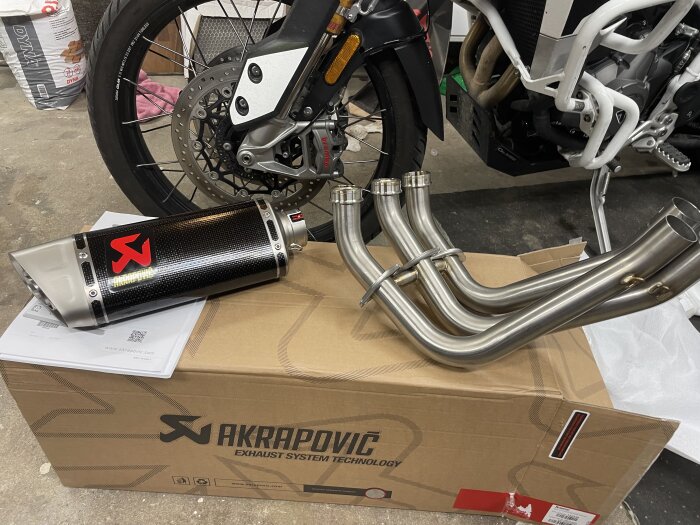 Exhaust system from Akrapovic shown next to a motorcycle wheel, placed on a cardboard box with Akrapovic branding in a garage.