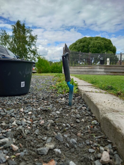 Närbild av en grusgång med en halvfylld hink och en planteringsspade i jorden, omgivna av grönska och byggnader i bakgrunden.