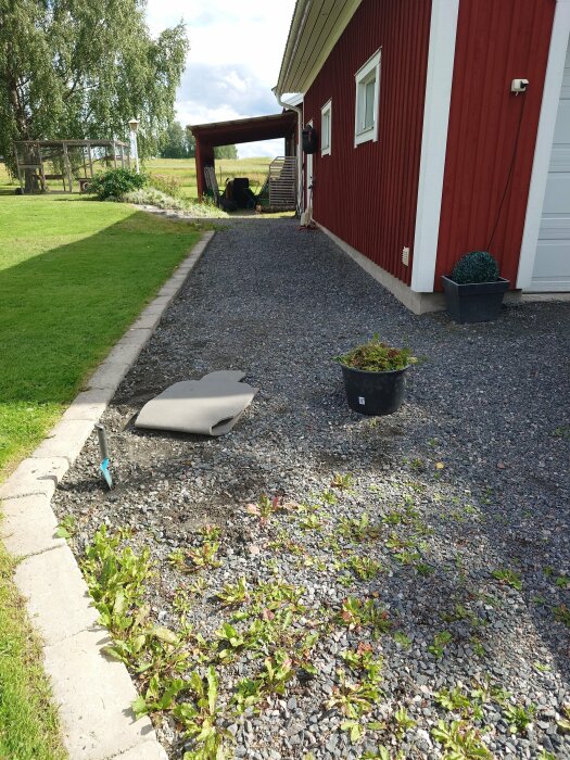 Grusgång med ogräs längs en röd byggnad, liggunderlag och verktyg syns. Växtlighet och redskap i bakgrunden. Klar blå himmel ovan.