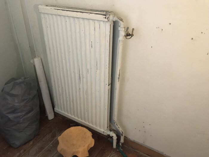 Gammal vit radiator, något rostig, monterad på en skadad vägg. Tygpåse och rör på vänstra sidan samt en gul trasa framför på trägolvet.