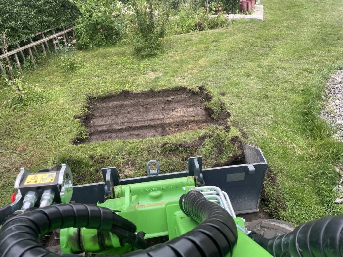 En grävmaskin skär rektangulära bitar av gräsmatta i en trädgård, omgiven av buskar och trädgårdsdekorationer.