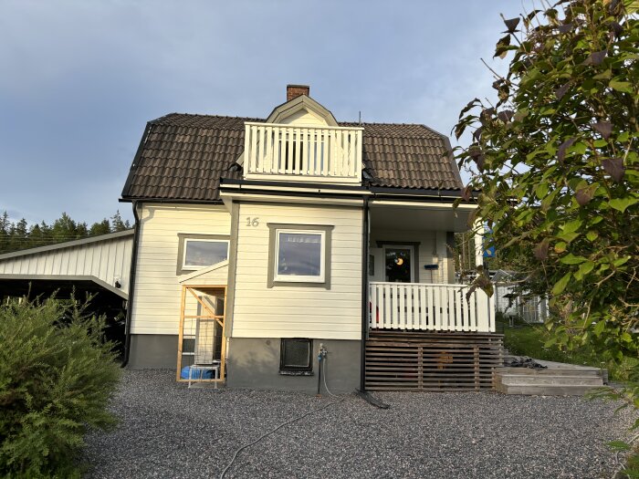 Vit tvåvåningshus med svart tak och balkong på andra våningen. Väderskydd under fönster och en stor buske till höger. Garagetaket syns till vänster.