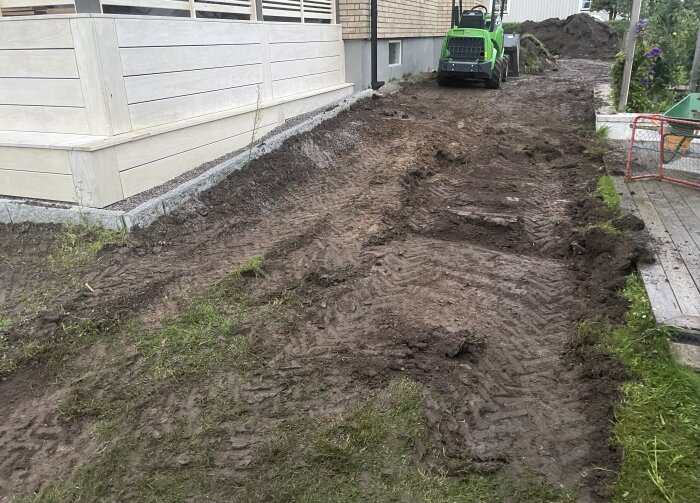 Grävd yta intill ett hus med lerigt underlag och ett grönt fordon i bakgrunden, efter att det börjat regna.