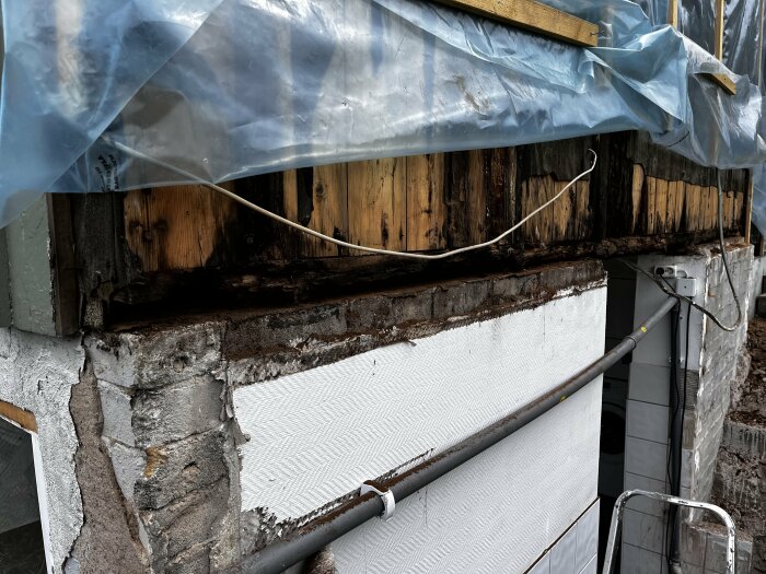 Skadad syll och fasad av ett hus med rötskador på träkonstruktionen ovanför grundmuren, samt plastskydd över delar av väggen.