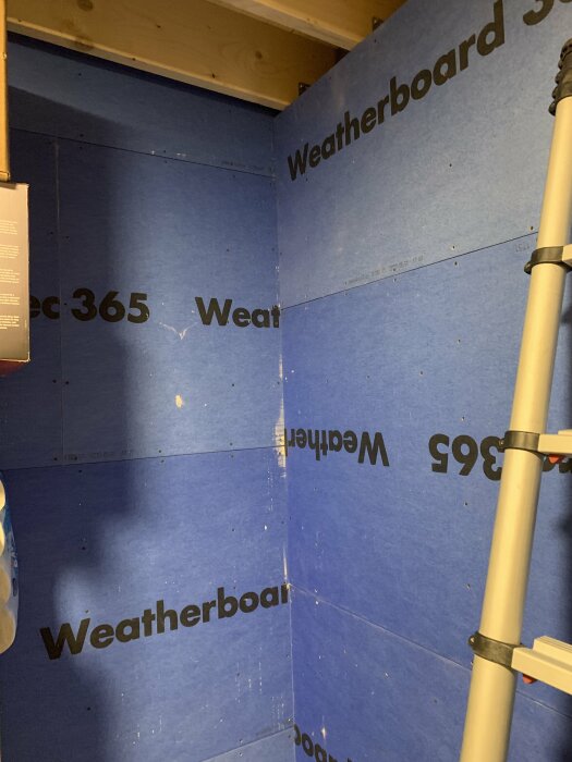 Väggar täckta med blåa gipsskivor märkt "Weatherboard 365", ingen isolering, synliga takbjälkar ovanför och en hopfällbar stege till höger i bilden.