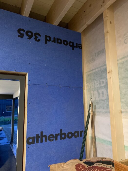 En vägg inomhus täckt med blåa gipsskivor märkta "Weatherboard 365" bredvid en vägg med plastfolie. Träbjälkar synliga i taket.