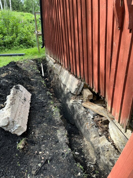 Röd ladugård med rätad och upplyft vägg. Marken runtom är uppgrävd och en bit av murstocken ligger på marken. Grönskande trädgård i bakgrunden.