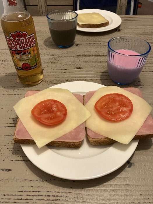 Två öppna smörgåsar med ost, skinka och tomat på en tallrik, Desperados-öl, samt glas med choklad- och rosa dryck på ett träbord.