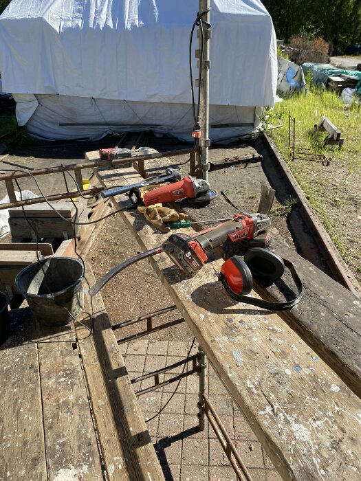 Verktyg och skyddsutrustning upplagda på en hakiställning utomhus, med en inplastad båt och diverse material i bakgrunden.
