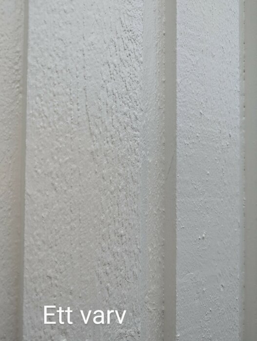 Bild av en husvägg målad med ett lager vit färg. Texten "Ett varv" syns på bilden. Ytan ser lite ojämn ut.