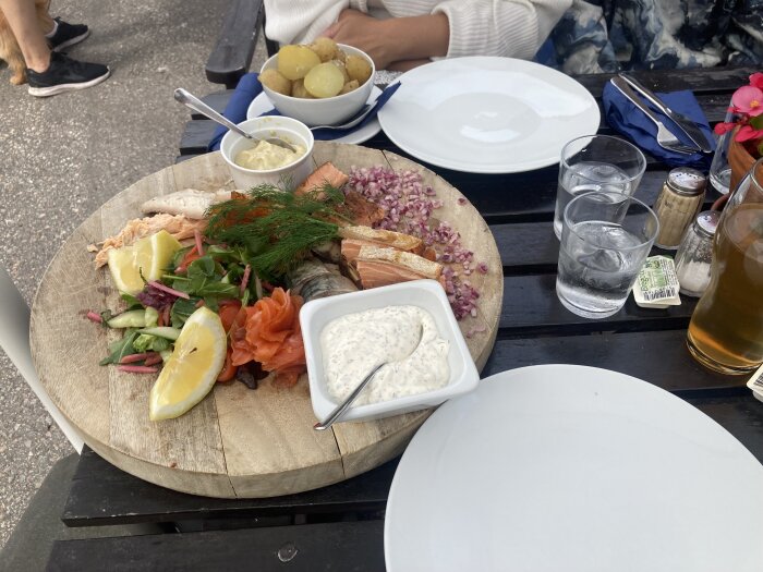 Lunch vid Håverud bestående av olika kallrökta och varmrökta delikatesser, serverad med potatis, sallad, citron och sås, på ett träfat utomhus.