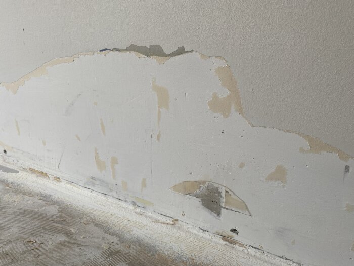Nedre delen av en vägg där målad tapet har börjat lossna, vilket avslöjar underliggande ytor som betong och gips.