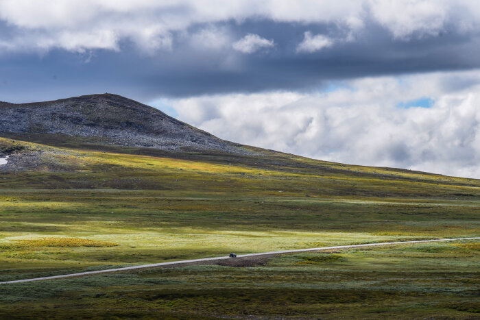 En ensam bil kör längs en väg genom ett stort, öppet landskap med gröna ängar och ett lågt berg i bakgrunden under en molnig himmel.