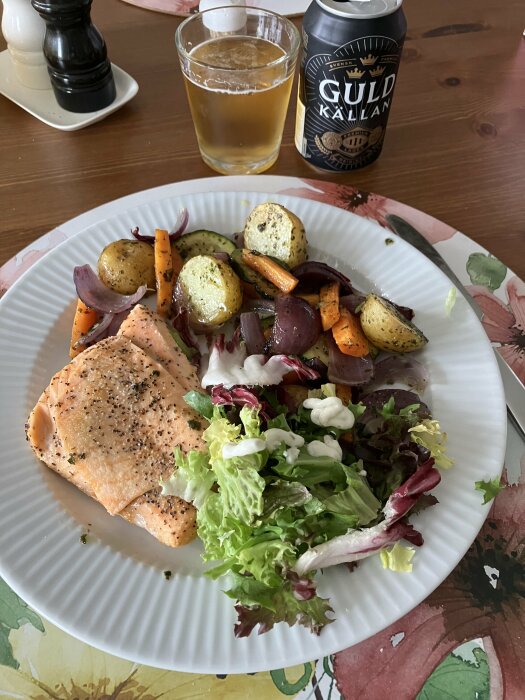 En portion ugnsbakad lax med rostade rotfrukter, blandad sallad med vitlökssås, samt en öl i glas och en ölburk på ett träbord.