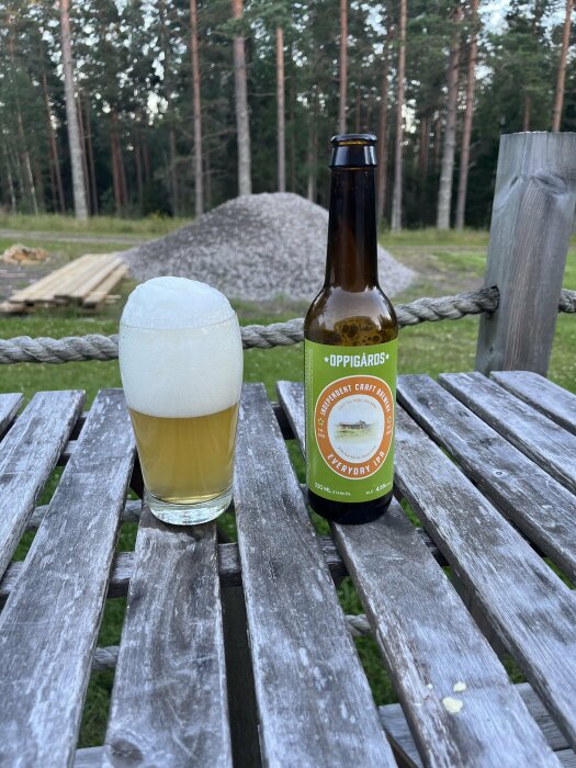 En flaska Oppigårds Everyday IPA och ett glas öl med skum på toppen står på ett slitet träbord utomhus, med en stor grushög och skog i bakgrunden.