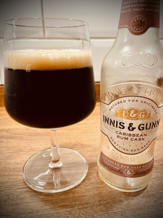 Ett glas mörkt öl med skumkrona bredvid en tom flaska Innis & Gunn Caribbean Rum Cask öl på ett träbord.