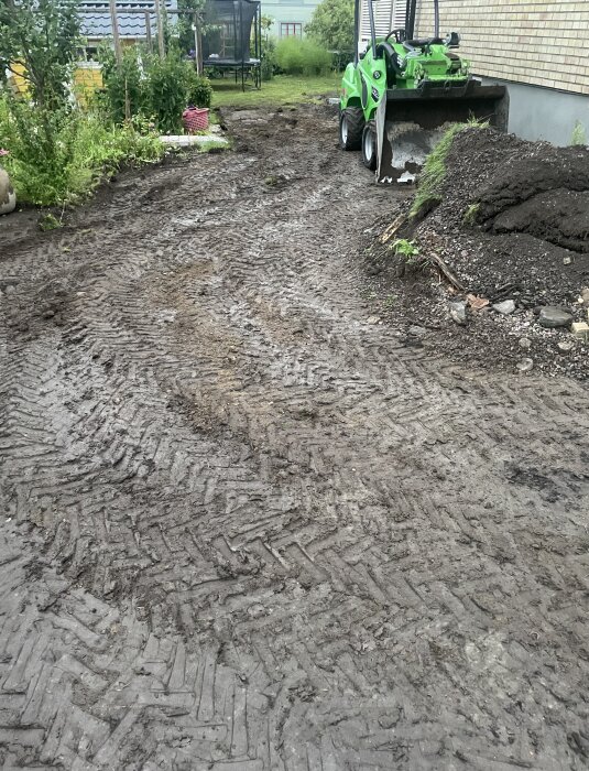 En lerig trädgård med traktorspår i marken efter mycket regn och en grävskopa från märket Avant parkerad bredvid en jordhög intill ett hus.