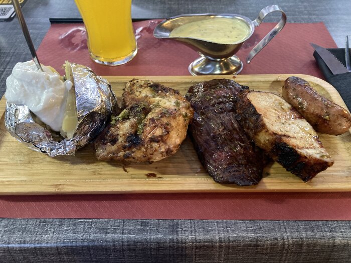 Mixed grill plate med bakad potatis, kyckling, kött, korv och sås på ett träfat med en öl i bakgrunden på restaurang.