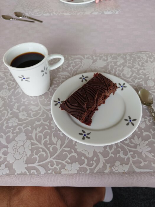 En kladdkaka på en vit tallrik med blommönster bredvid en kopp svart kaffe på ett bord med mönstrad duk.