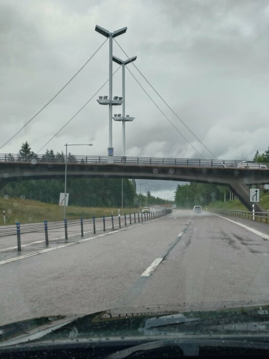 En bil kör på en regnig väg under en bro med höga lyktstolpar och vajrar. Skog och vägskyltar syns i bakgrunden.