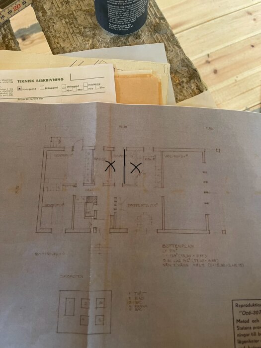 Ritning över bottenplan av ett hus med texter och markeringar som föreslår ändringar i planlösningen. Ritningen ligger på ett träbord med tekniska dokument ovanpå .