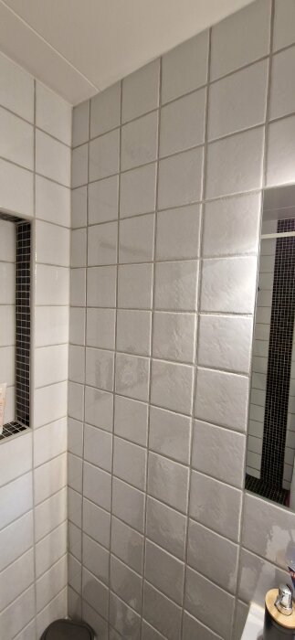 Vägg i badrum täckt med vita kakelplattor, spegel till höger och hylla inbyggd i väggen till vänster med små svarta mosaikplattor.