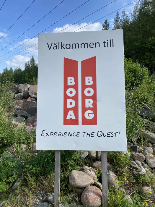 Vägskylt med texten "Välkommen till Boda Borg, Experience The Quest!" i naturmiljö med stenar och växtlighet runtom.