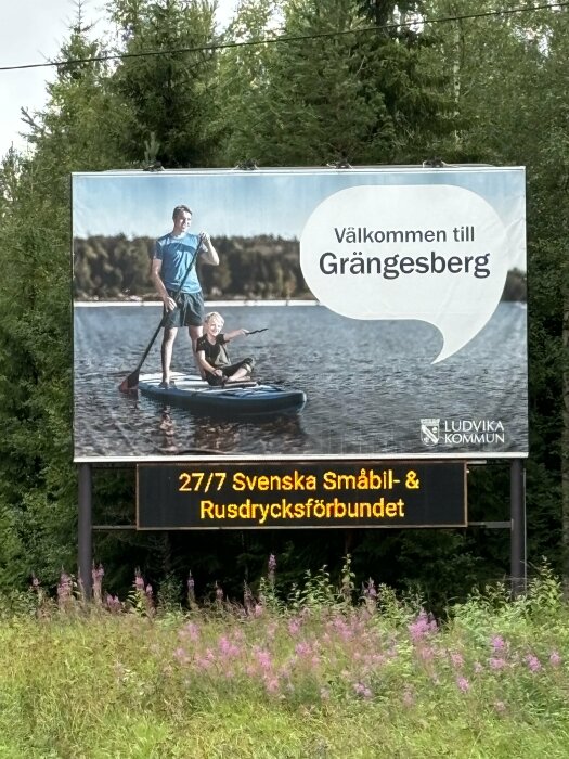 Vägskylt med text "Välkommen till Grängesberg" och foto av man och barn på SUP-bräda på vatten, samt digital text om evenemang för Svenska Småbil- och Rusdrycksförbundet.