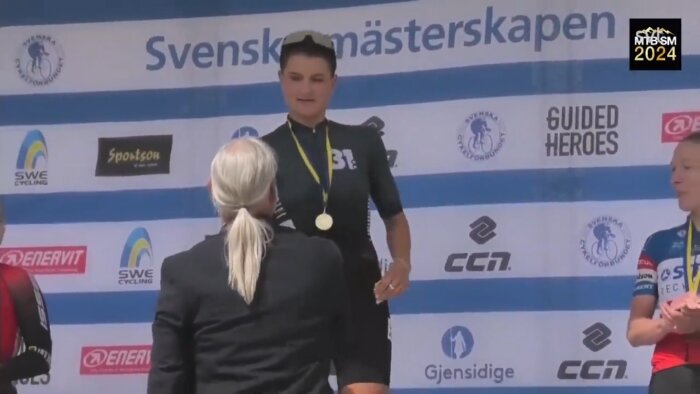 En person med medalj står på prispallen vid Svensk mästerskapstävling, gratulerad av en annan person, framför en sponsorskylt.