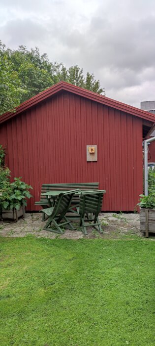 Grön trädgårdsmöbelgrupp framför en röd stuga med fågelholk på väggen, omgiven av grönska under en molnig himmel.