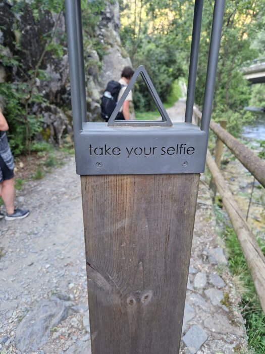 Trästolpe med metallram och texten "take your selfie" på en grusväg i skogen, två personer syns vandrandes i bakgrunden.