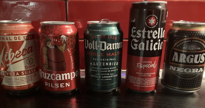 Fem burkar med olika ölsorter: Xibeca Damm, Cruzcampo Pilsen, Voll-Damm Doble Malta, Estrella Galicia och Argus Negra, placerade på en mörk yta mot en röd bakgrund.