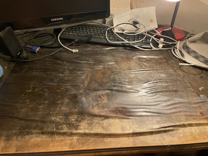 Gamla skrivbordets fanér skadat av vatten med upphöjningar och repor, med dator, tangentbord, kablar och hatt på bordet.