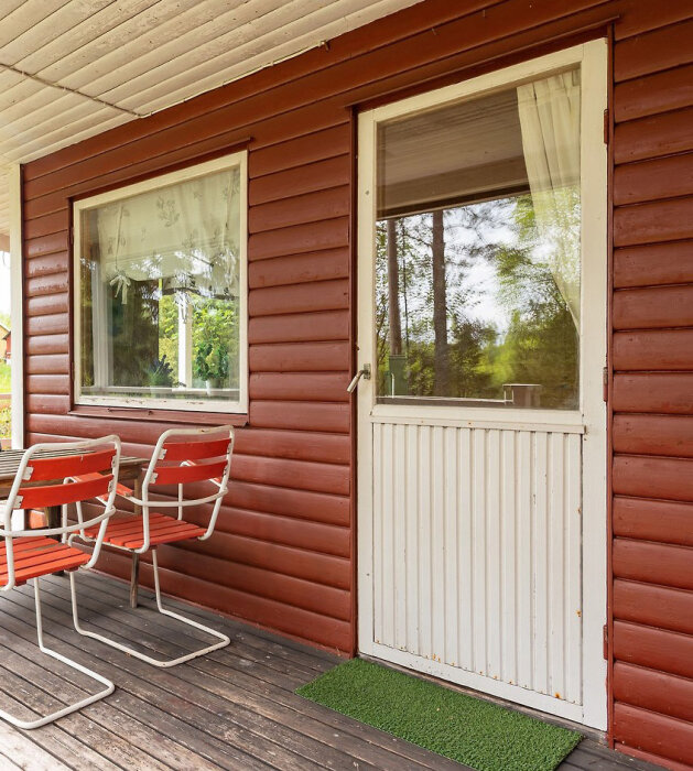 Röd sommarstuga med vita enkelglasfönster och en vit dörr utan foder. Två rödvita stolar vid ett bord står på verandan framför.