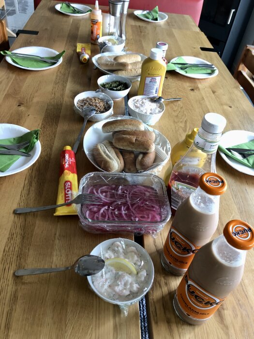 Middagsbord med korvbröd, bröd, senap, ketchup, picklad rödlök, räksallad, friterad lök, chokladmjölk och papptallrikar med gröna servetter.