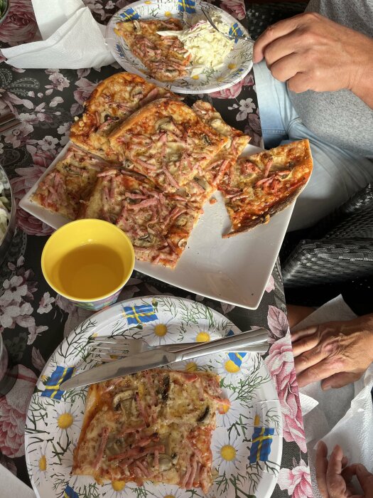 Hemlagad pizza med skinka, lök och ost, serverad på blommiga papperstallrikar tillsammans med pizzasallad. En gul kopp och personer som äter syns också.