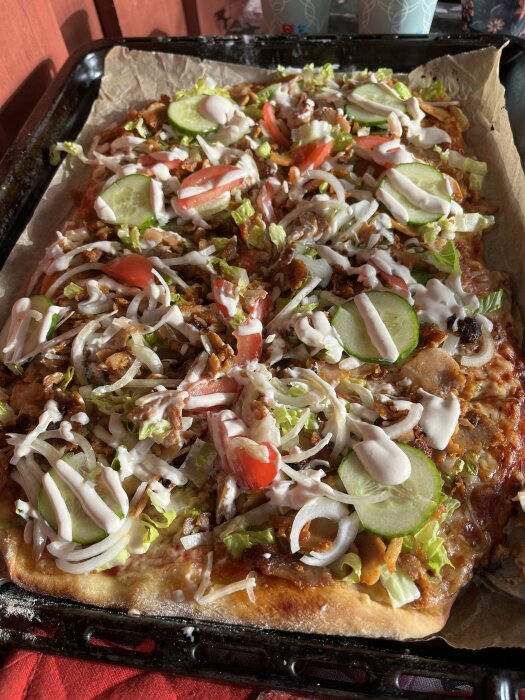 En hemmagjord rektangulär pizza med kycklingkebab, sallad, gurka, tomat, ost, lök och kebabsås på en plåt med bakplåtspapper.