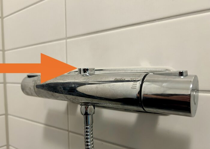Närbild på en kromad duschblandare med en orange pil som pekar på en specifik punkt där avståndet från blandaren till väggfästet har mätts.