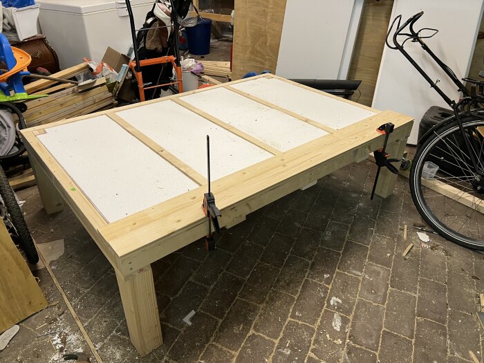 Hemmagjort arbetsbord med plywood och frigolitisolering, fäst vid kanterna med träskruvar, står i en verkstad med verktyg och cykel runtomkring.