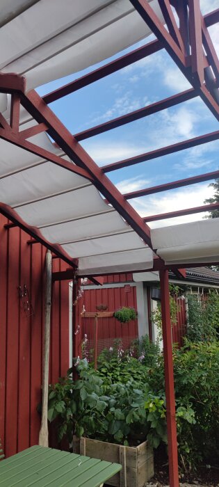 Pergola med vitt tyg uppsatt, bredvid en röd träfasad, grönskande trädgård med planterade växter och en grön bordsyta i förgrunden.