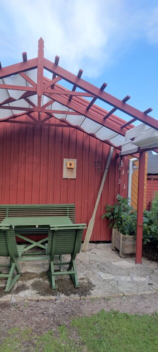 Pergola med duk över matplats utomhus framför röd byggnad. Grönt bord med tre stolar, en kratta står lutad mot väggen vid en blomkruka.