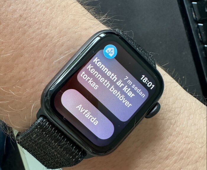 En smartwatch på en handled visar notisen "Kenneth är klar, Kenneth behöver torkas" från en Home Connect-app. Tid på klockan är 18:01.