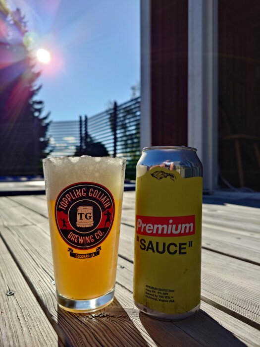 En burk med öl märkt "Premium Sauce" och ett fyllt glas med Toppling Goliath Brewing Co-logga, placerade utomhus i solljus på trädäck.