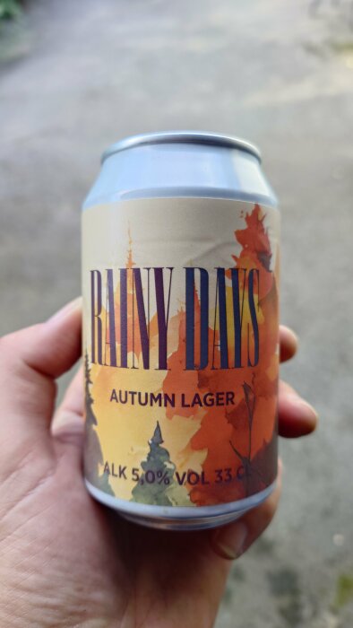 En hand håller i en burk med öl märkt "Rainy Days Autumn Lager" med höstlöv i bakgrunden.